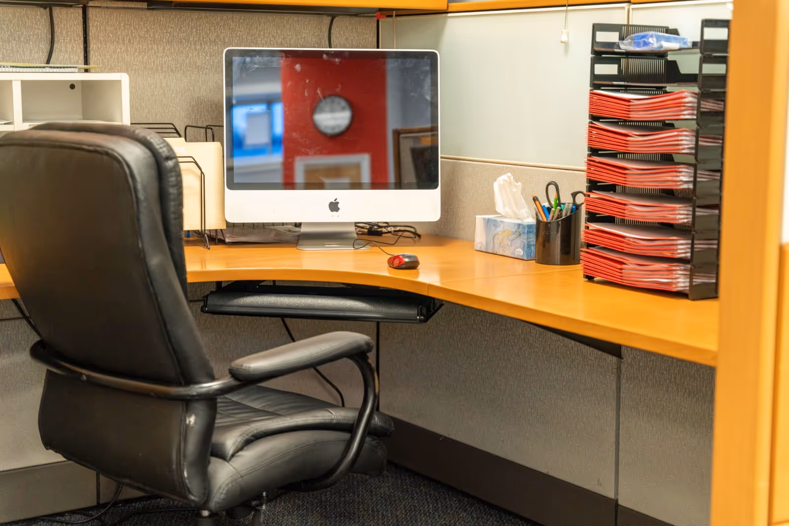 Office cubicle with an iMac computer, black leather chair, tissue box, and red file organizers.