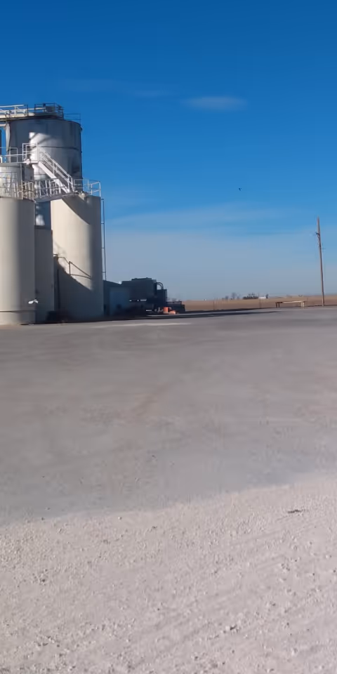 Industrial silos and equipment sit on a wide, open, paved outdoor area under a blue sky.