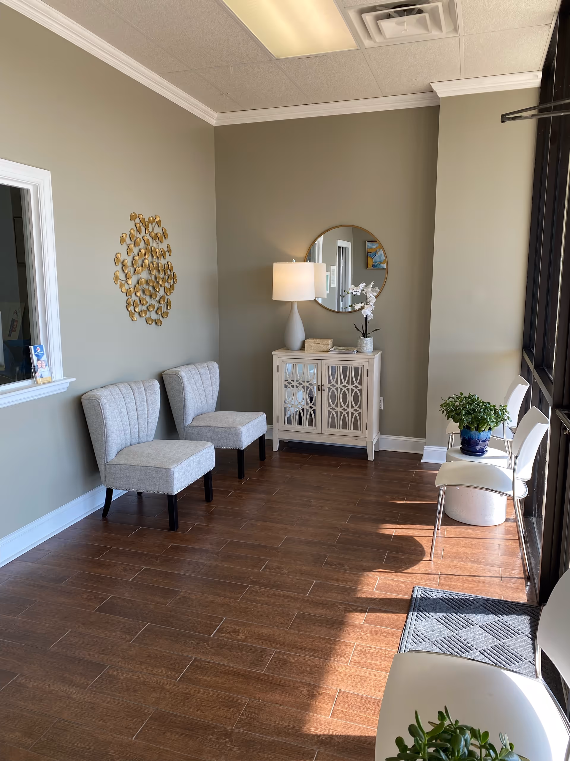 Interior waiting area with gray chairs, a console cabinet, mirror, and plants.