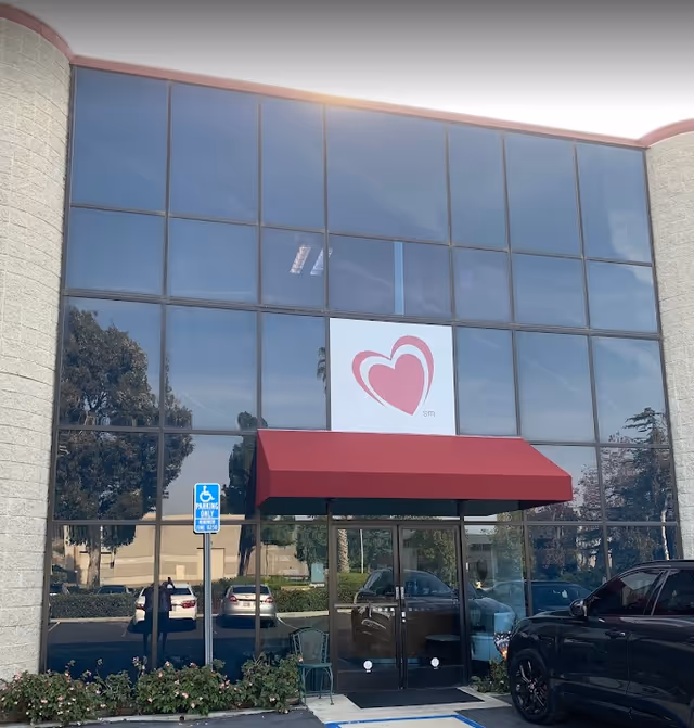 Glass-front entrance of a senior living facility with a red awning and heart logo.