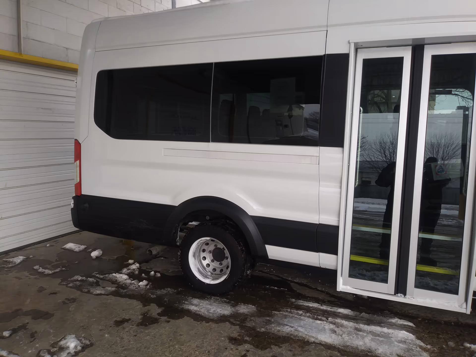 White mobility shuttle van parked beside a building with a large garage door, snow on ground.