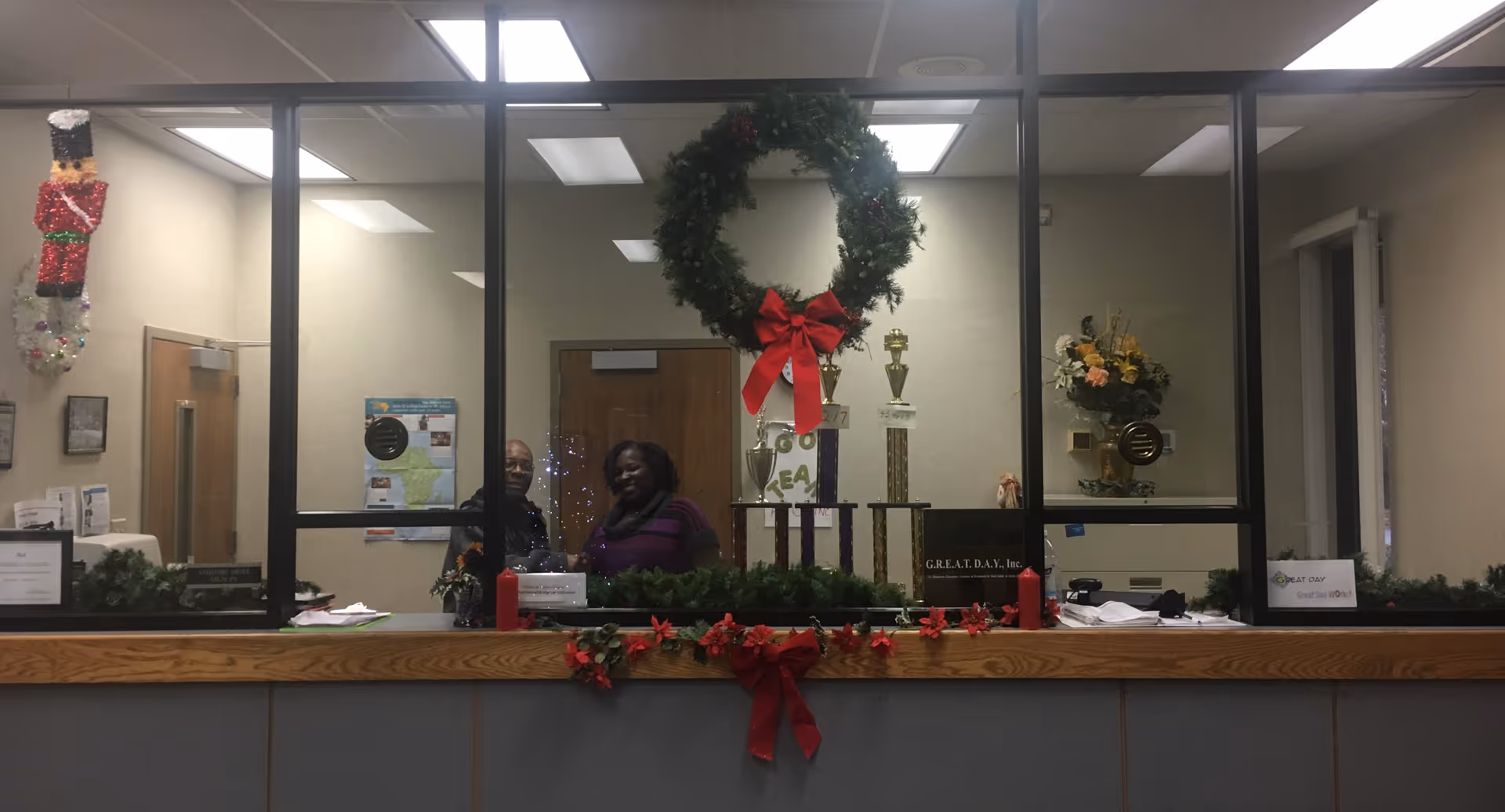 Indoor reception area with festive wreaths, garland, and two staff behind a glass partition.