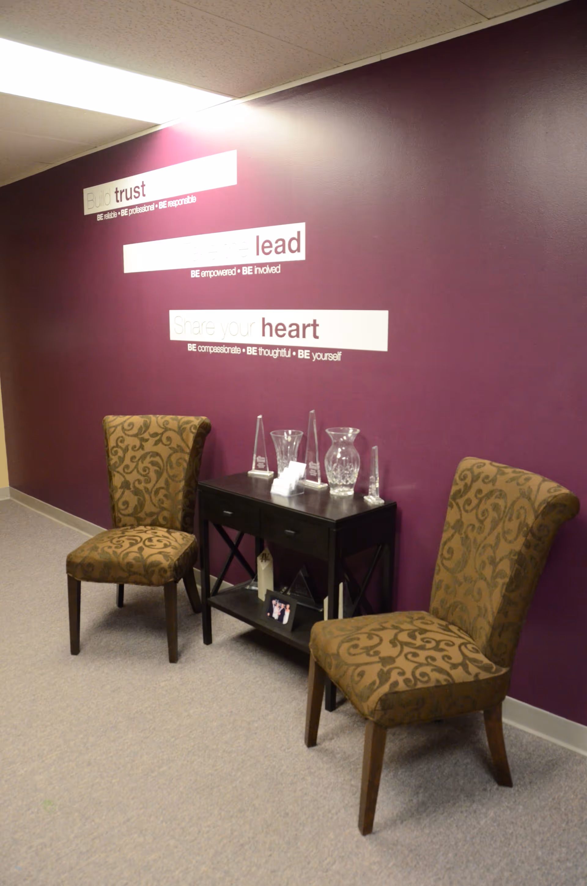Two patterned chairs flank a black console table against a purple wall with inspirational quotes.