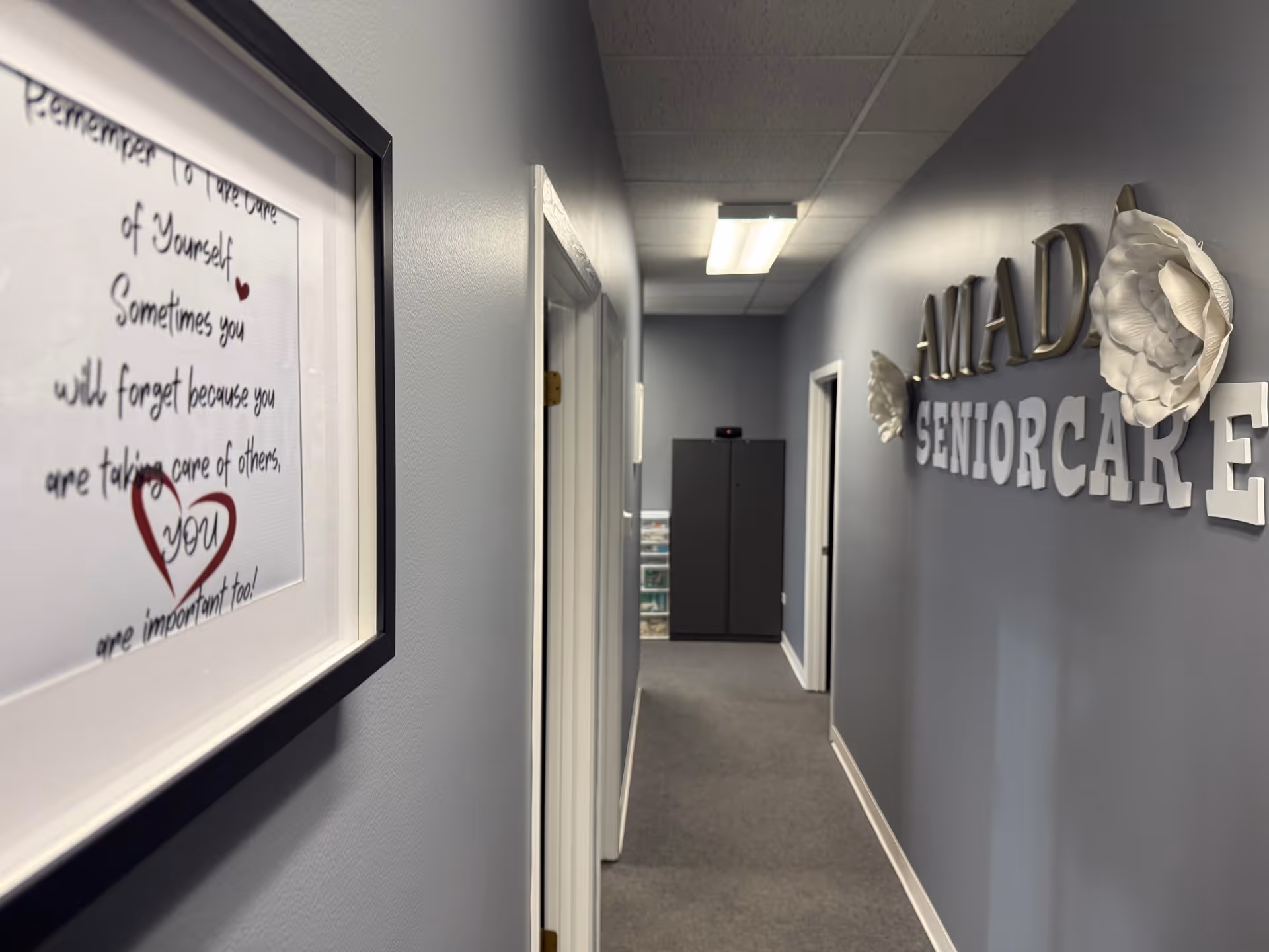 Inside a quiet Amada Senior Care hallway featuring wall signage, framed art, and a gray corridor.