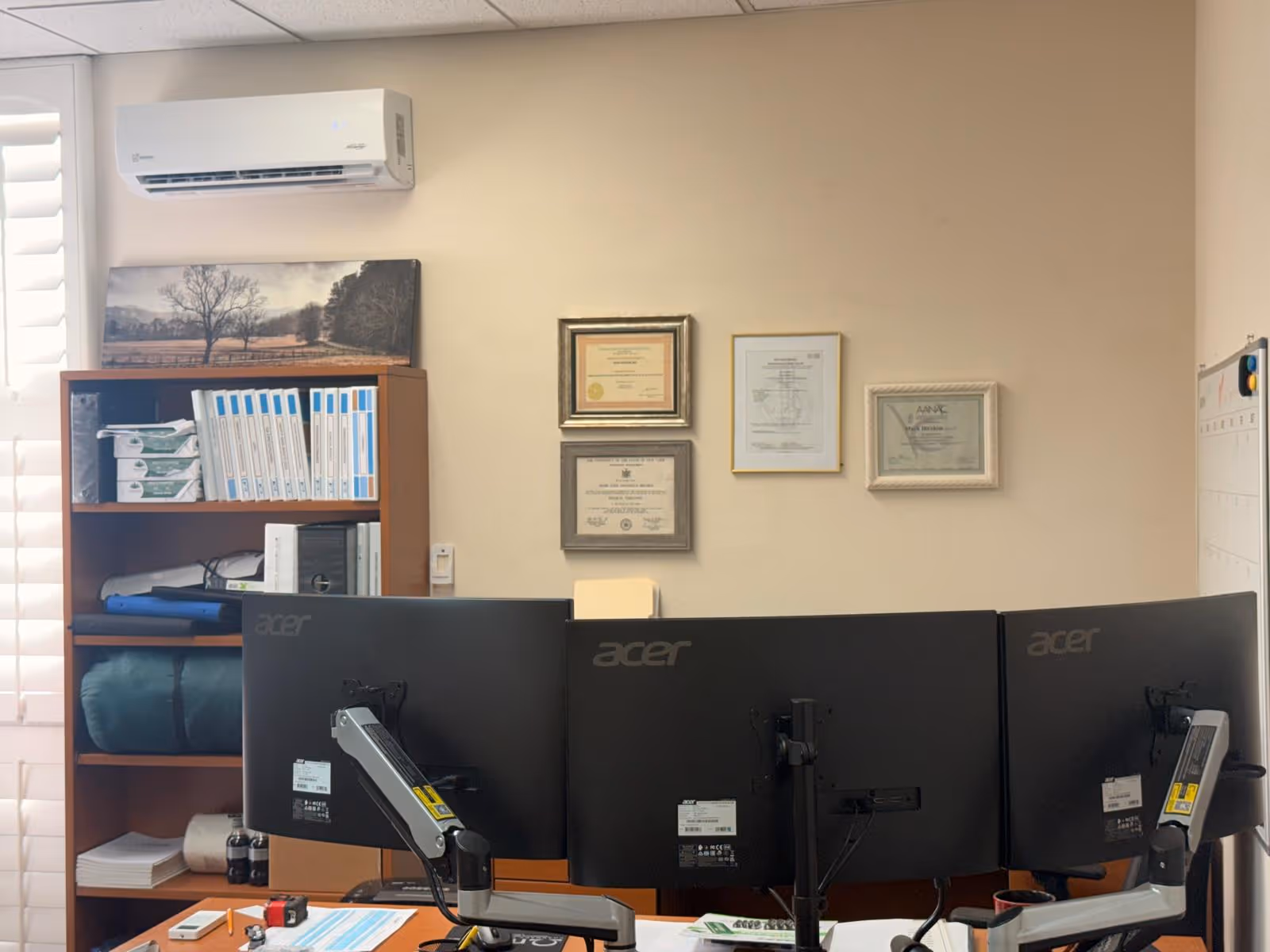 Office workstation with three Acer monitors, shelves, and framed certificates on a beige wall