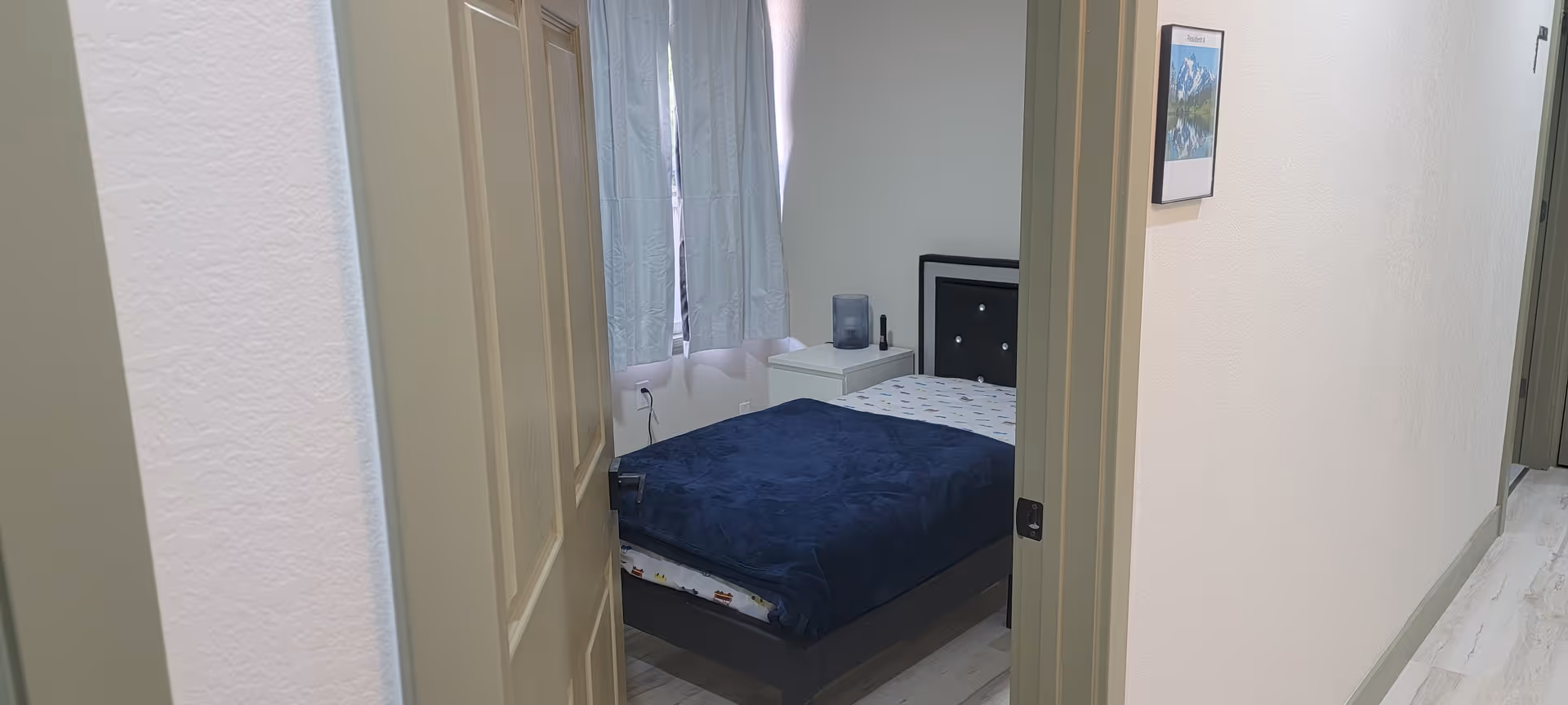 A bedroom in a senior living facility with a bed, nightstand, and window curtains.