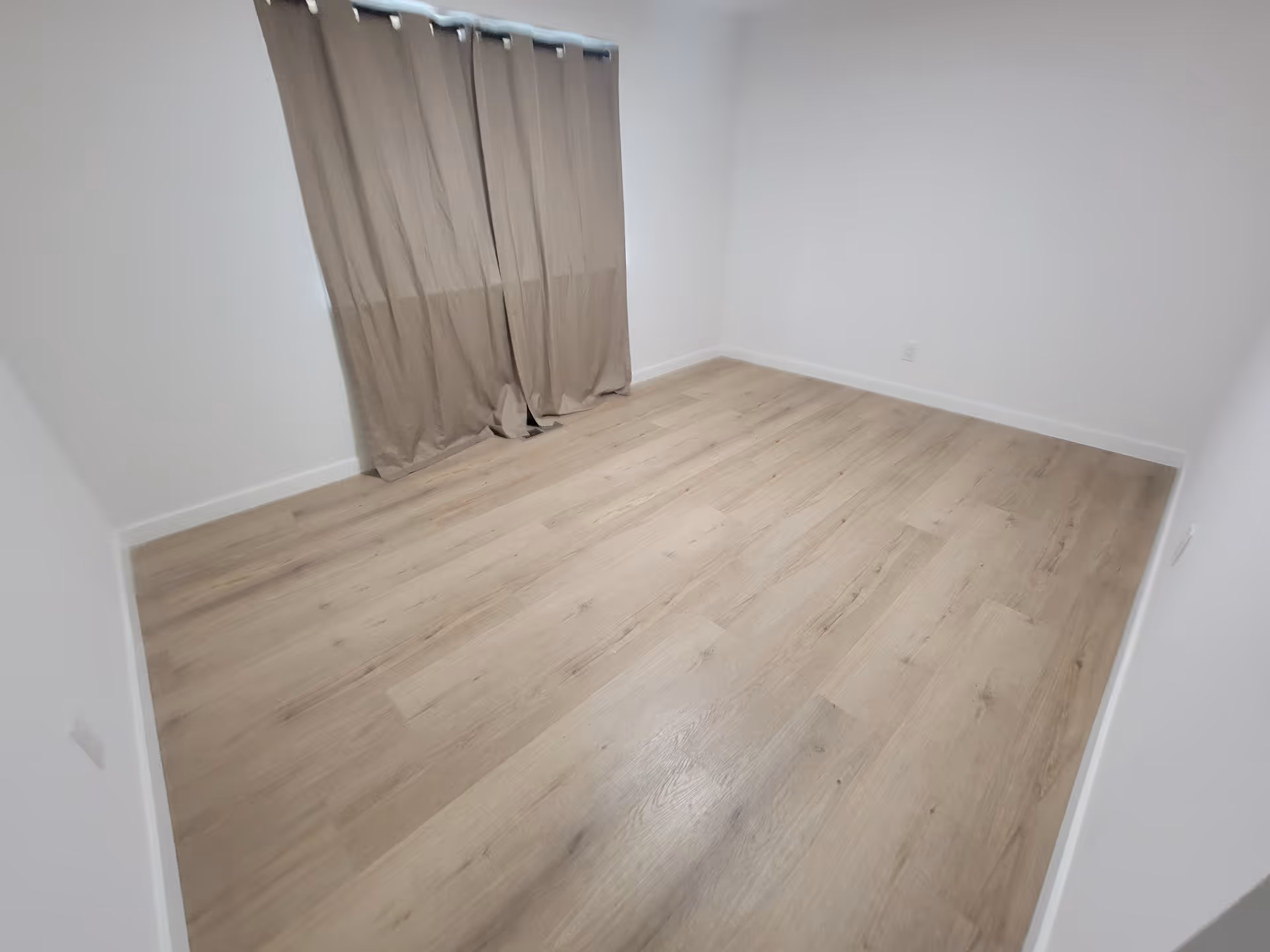 An empty room with light wood flooring and beige curtains covering a window.