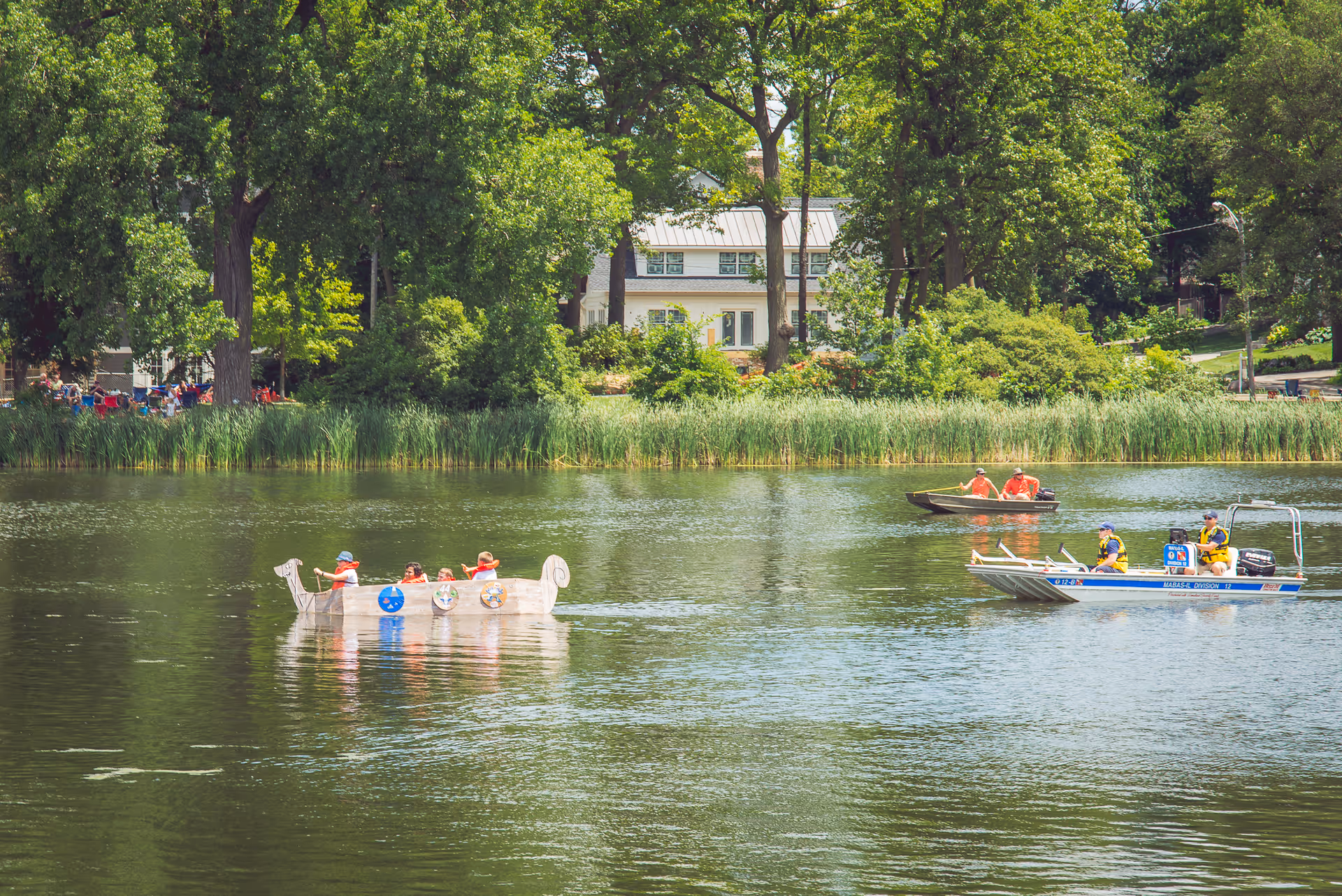 Outdoor lake scene with people in small boats and a tree-lined campus backdrop.