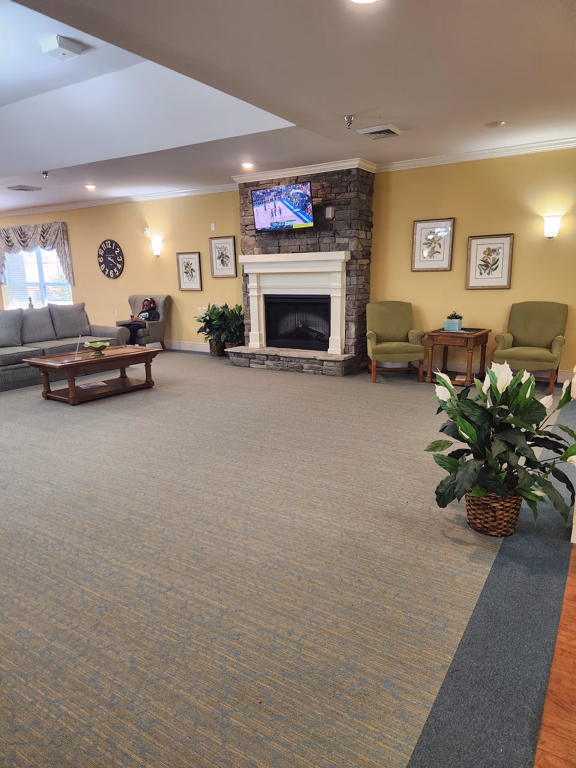 Senior living lobby featuring stone fireplace, TV, beige walls, sofas, chairs, and potted plants.