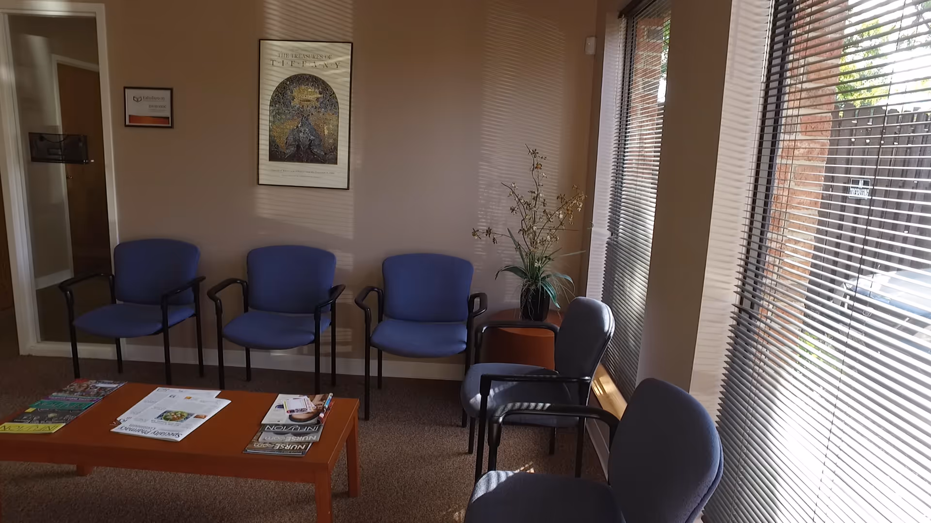 Indoor waiting area with blue chairs, magazines, a coffee table, and large window blinds.
