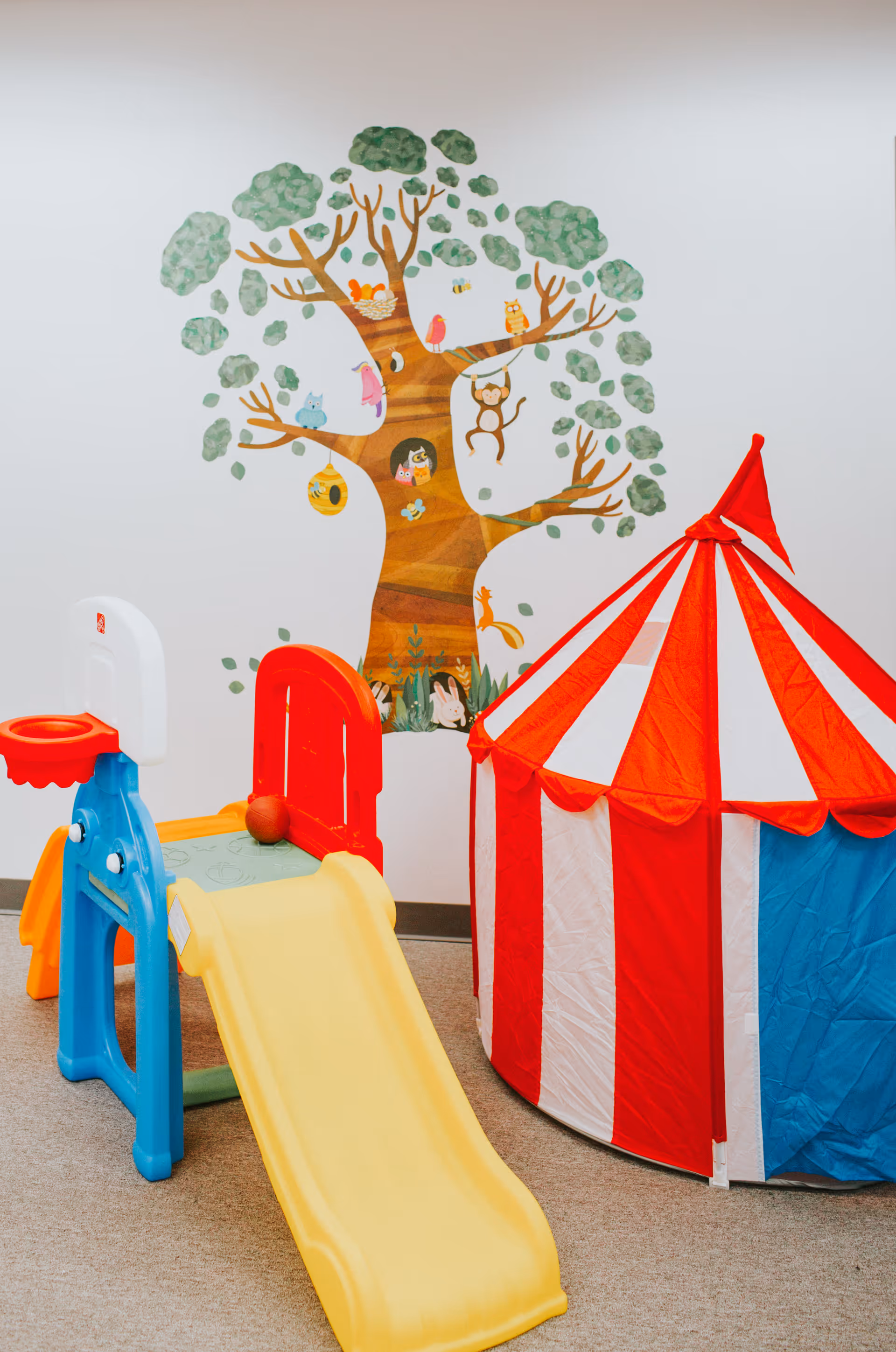Interior children's play area with a yellow slide, red-and-white tent, and tree mural.