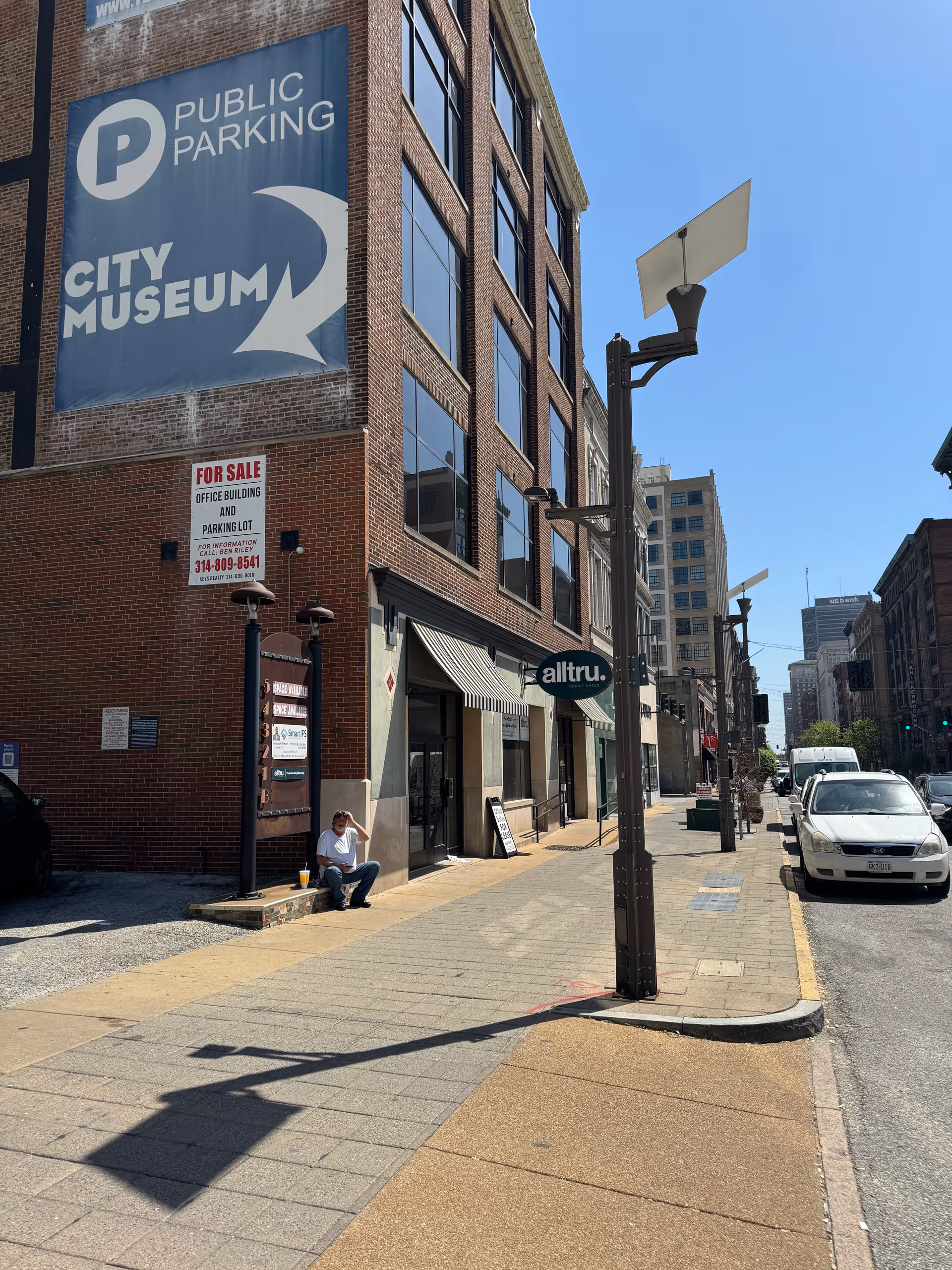 City street scene with brick building, large signage, storefronts, and a man sitting on the curb.
