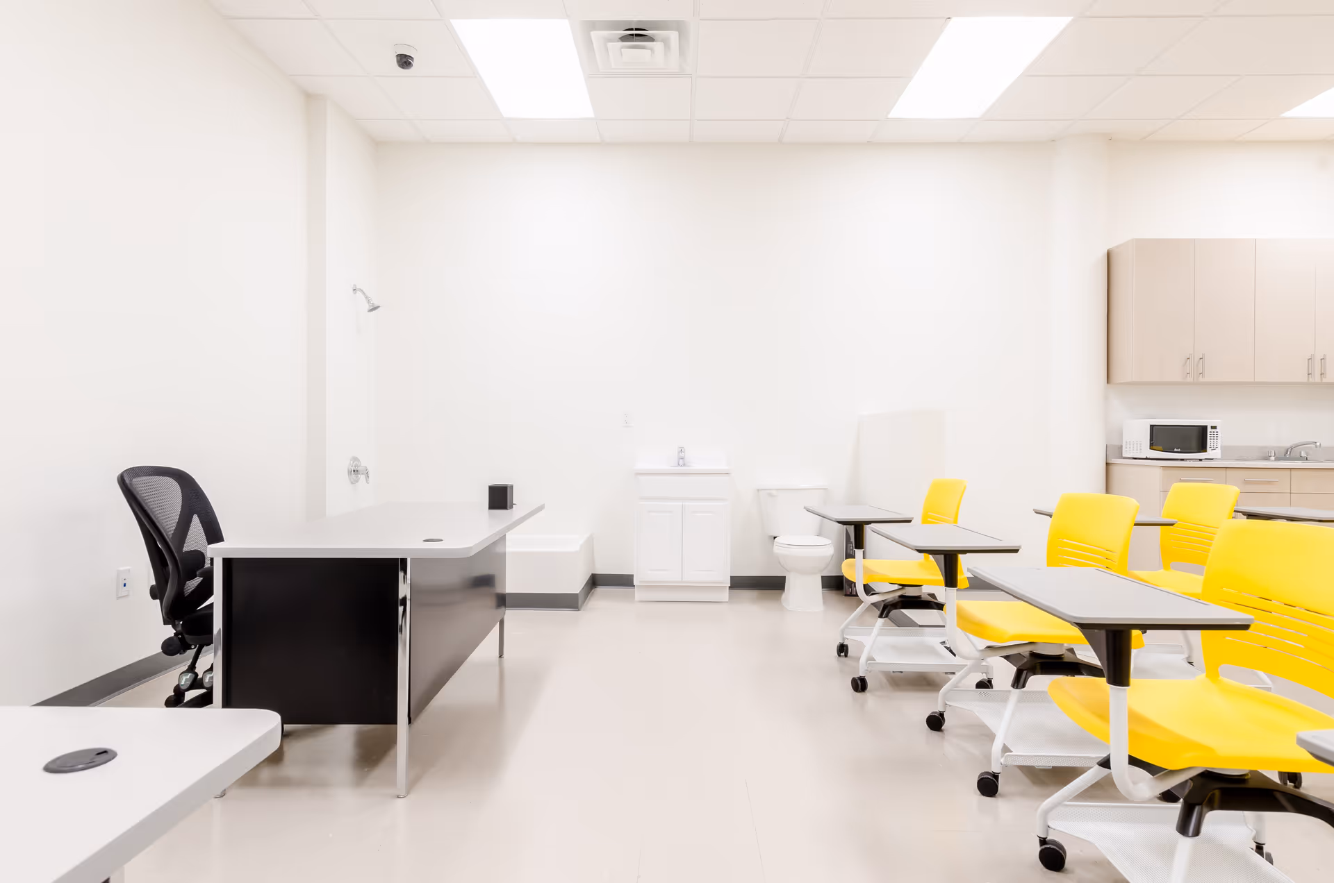 Bright institutional room with yellow mobile desks, a black desk, kitchenette, sink, and toilet.