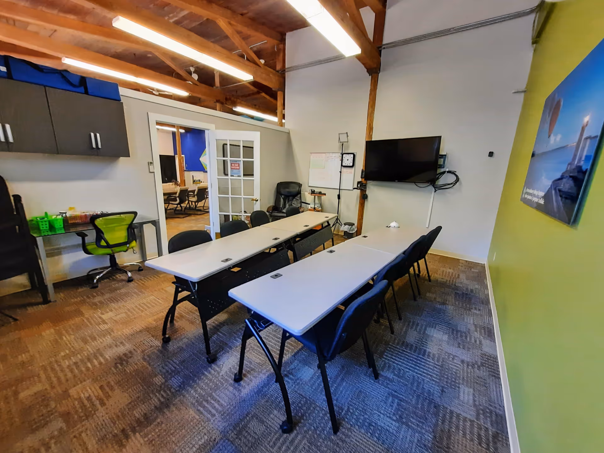 A conference-style meeting room with tables, chairs, a wall-mounted TV, and a doorway to another area.