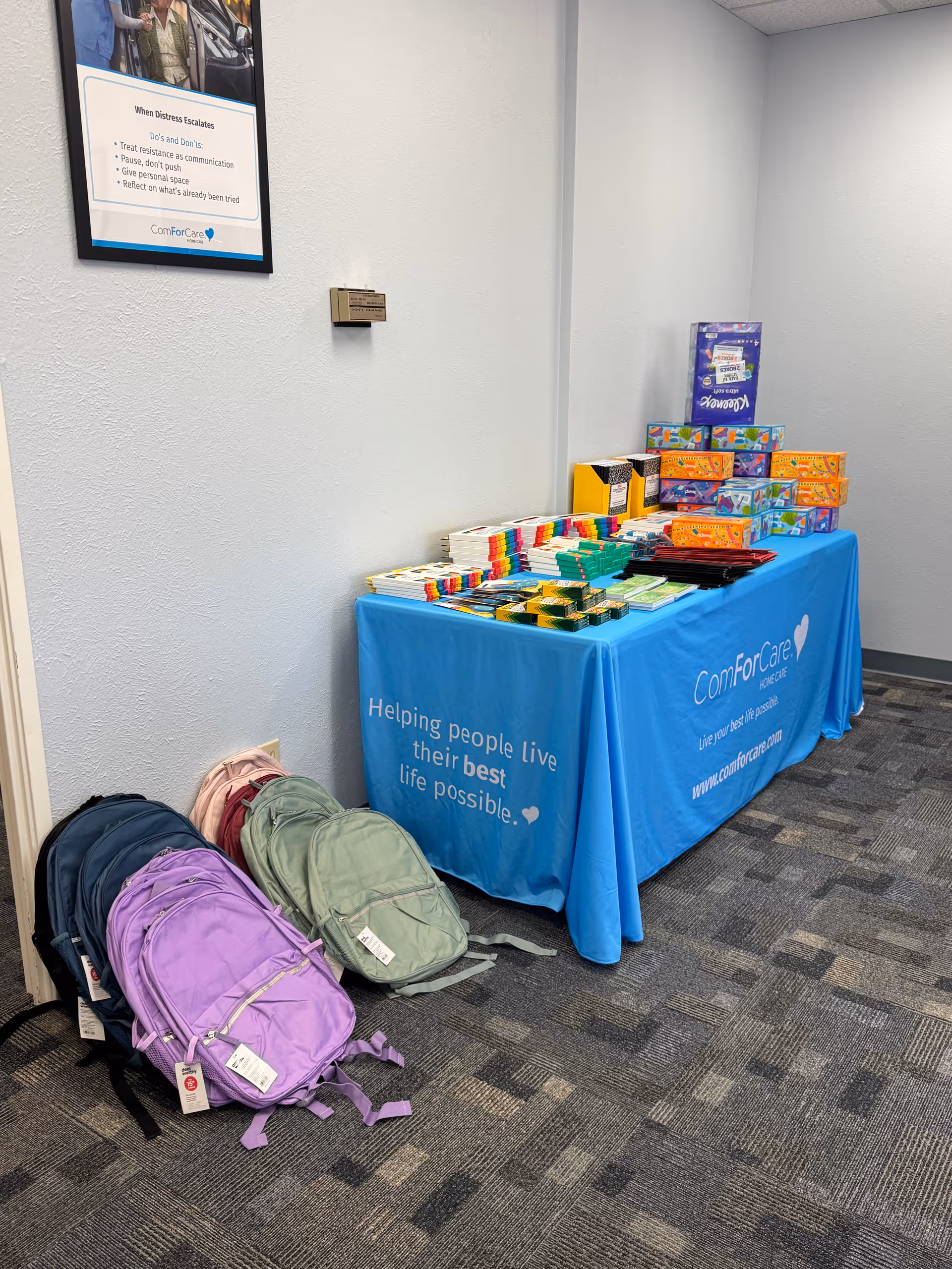 An indoor information table with a blue ComForCare tablecloth, supplies, and backpacks.