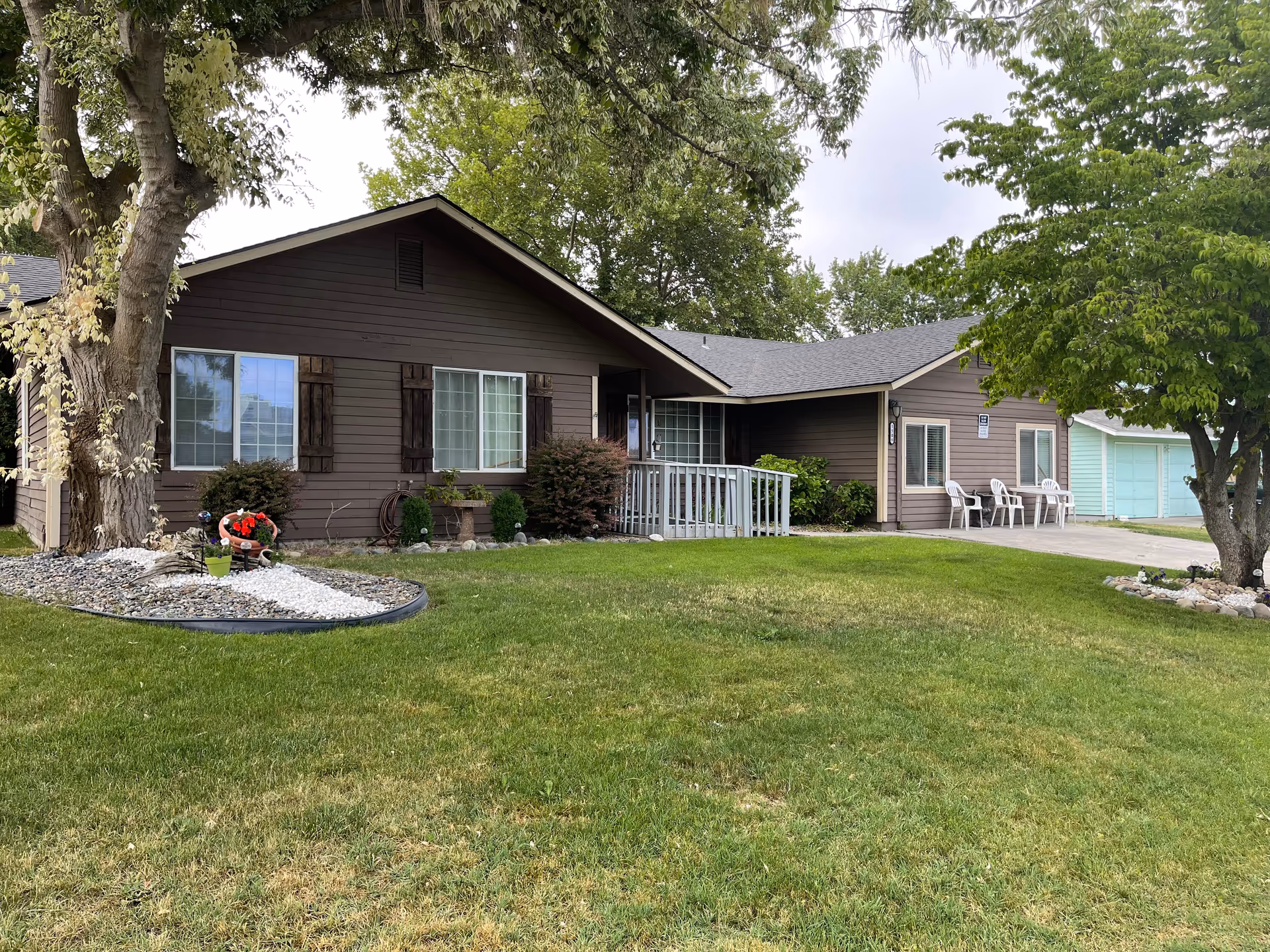 Single-story brown house with a manicured front lawn, trees, and white porch chairs.