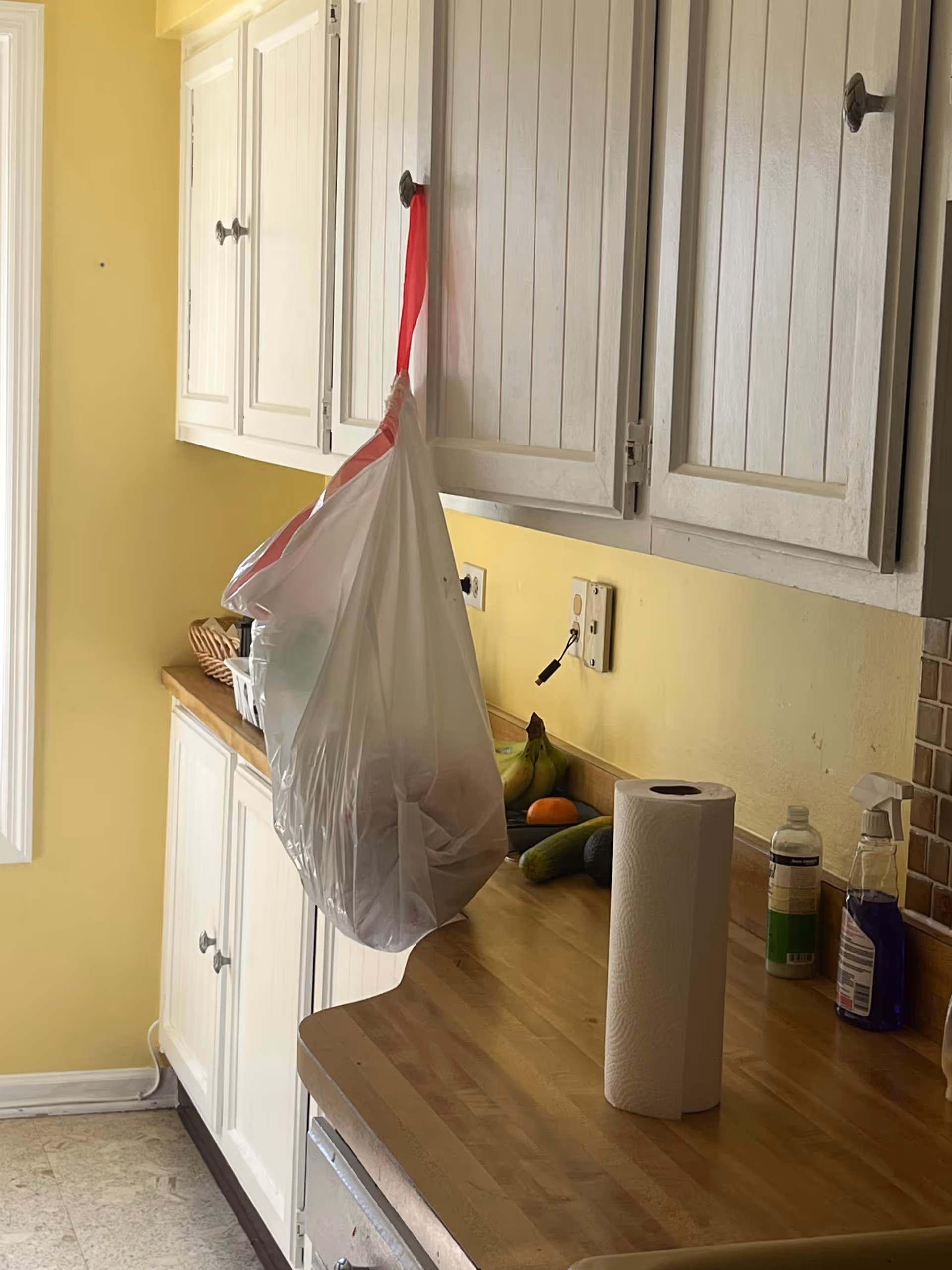 Plastic bag hangs from cabinet over a yellow kitchen counter with fruit.