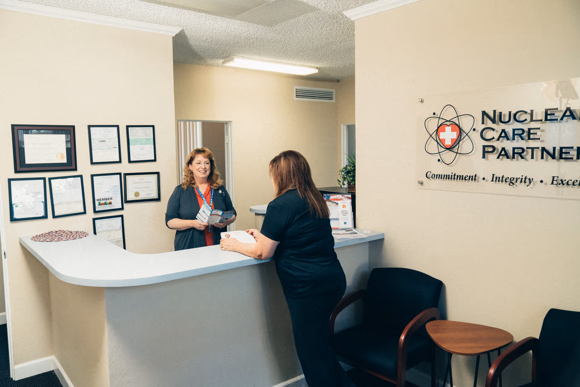 Two staff members greet a resident at a reception desk inside a Nuclear Care Partners lobby.