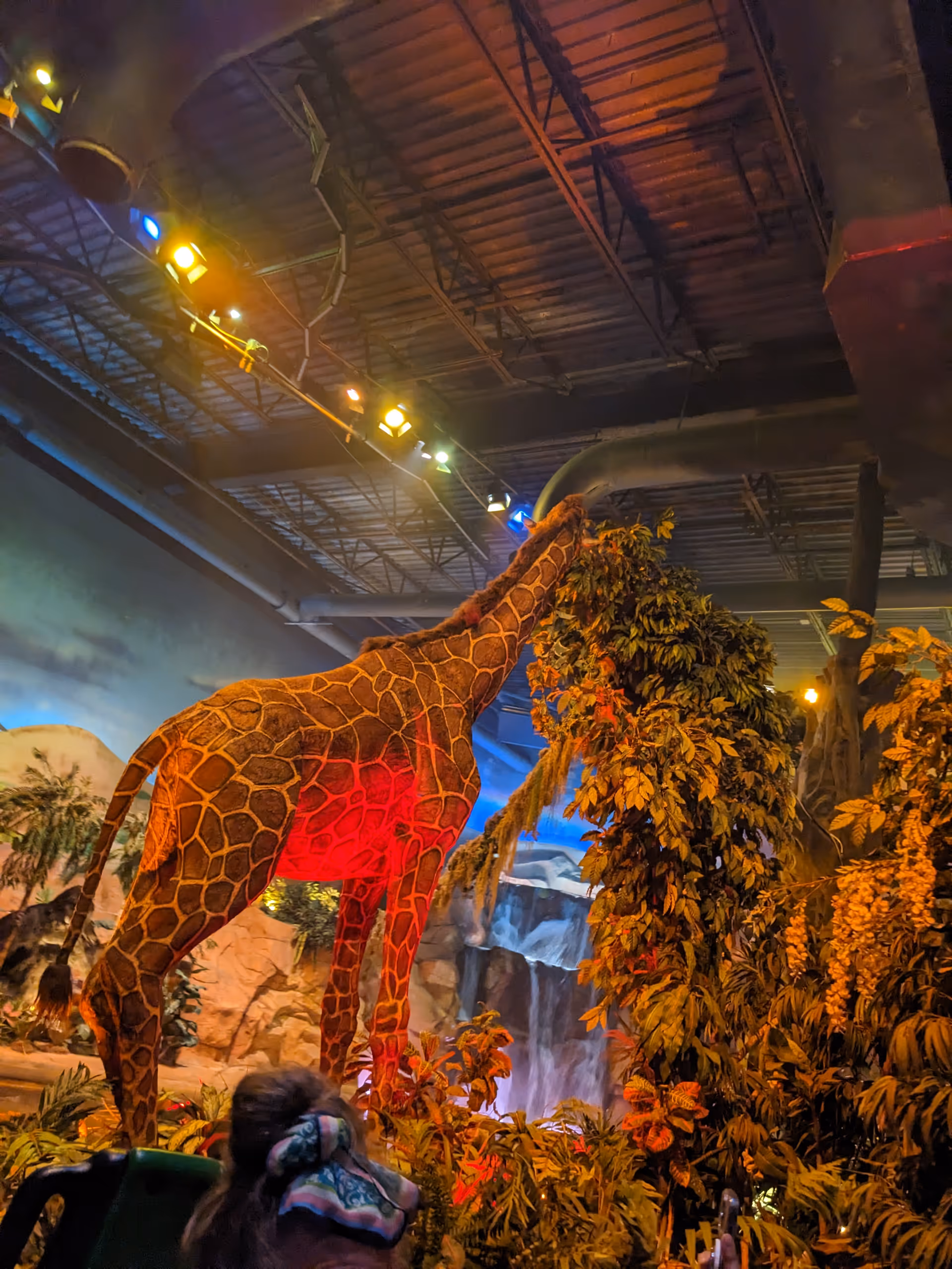 Indoor exhibit with a life-sized giraffe statue, jungle plants, and colorful lighting.