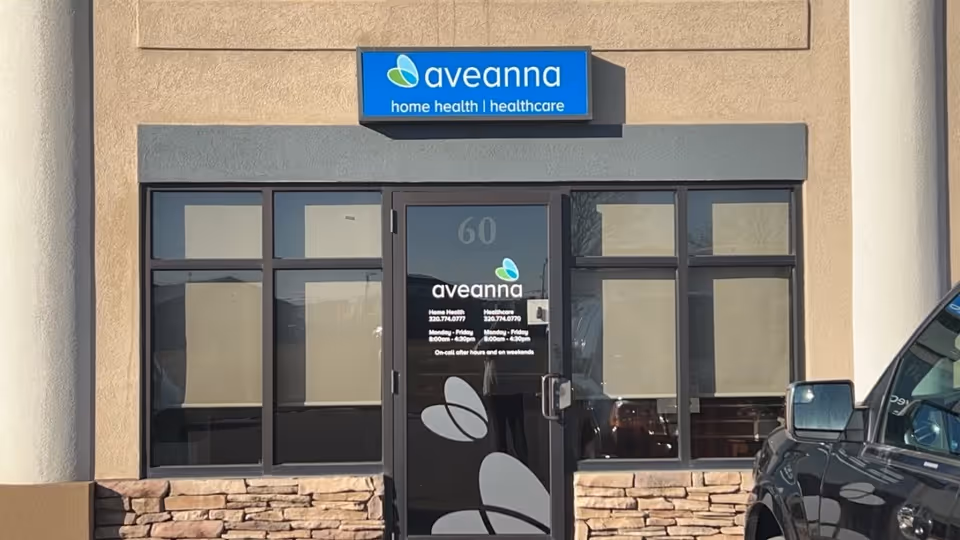 Front entrance of Aveanna Home Health building with glass doors and parked vehicle.