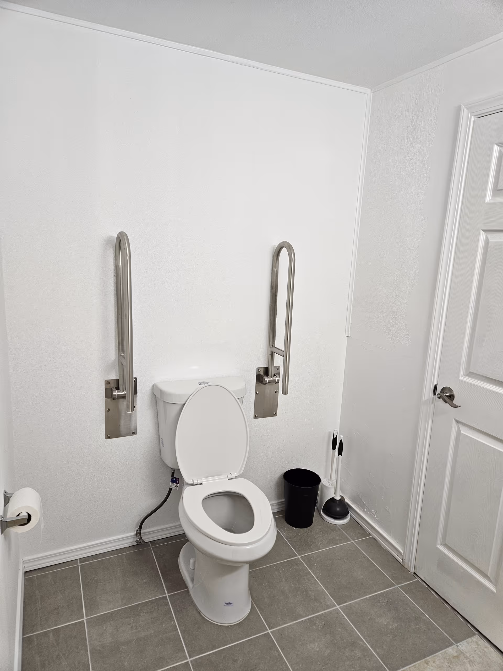 Accessible bathroom with toilet, support rails, and cleaning supplies near a door.