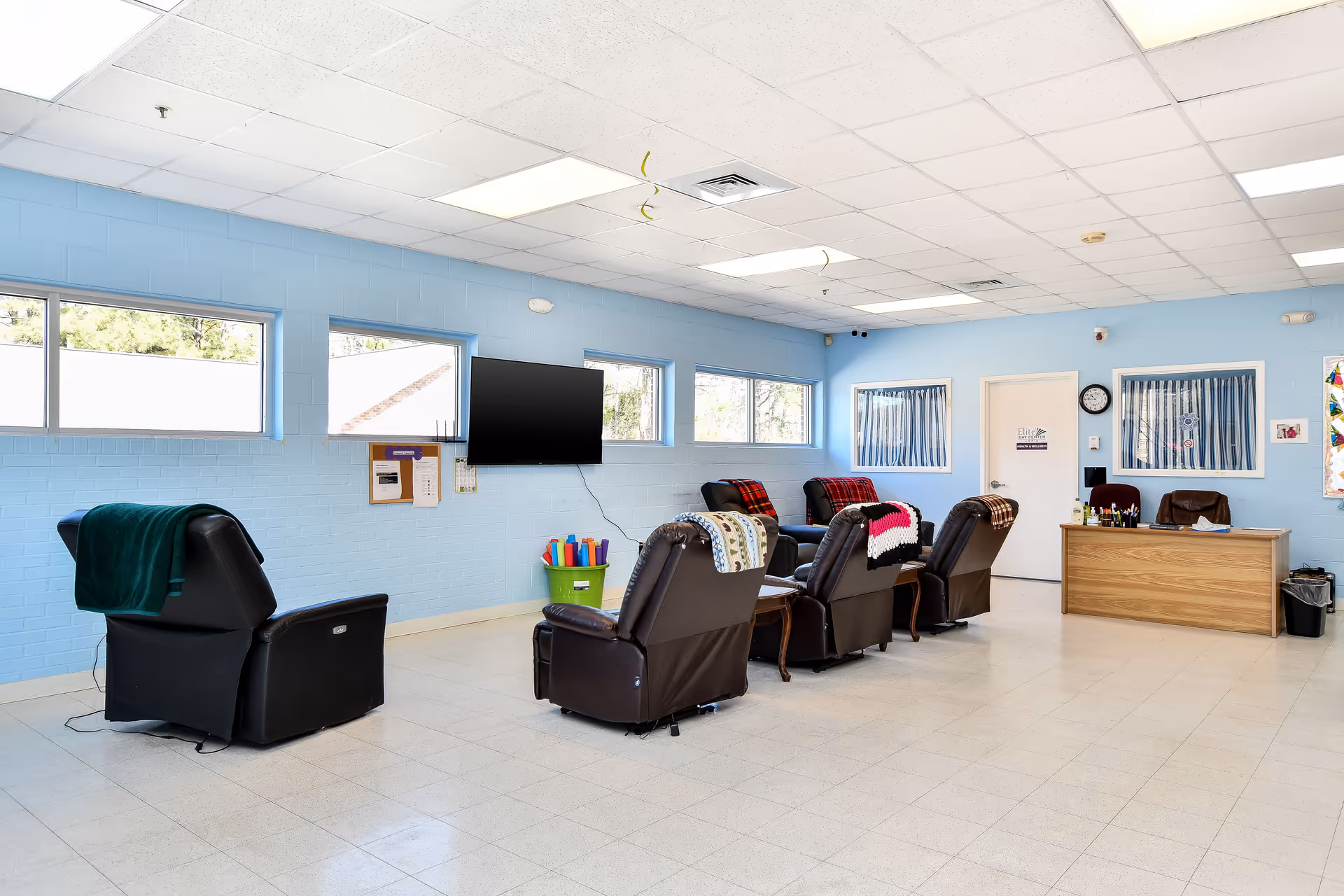Bright blue-walled senior lounge with multiple recliners, a wall-mounted TV, and a reception desk.