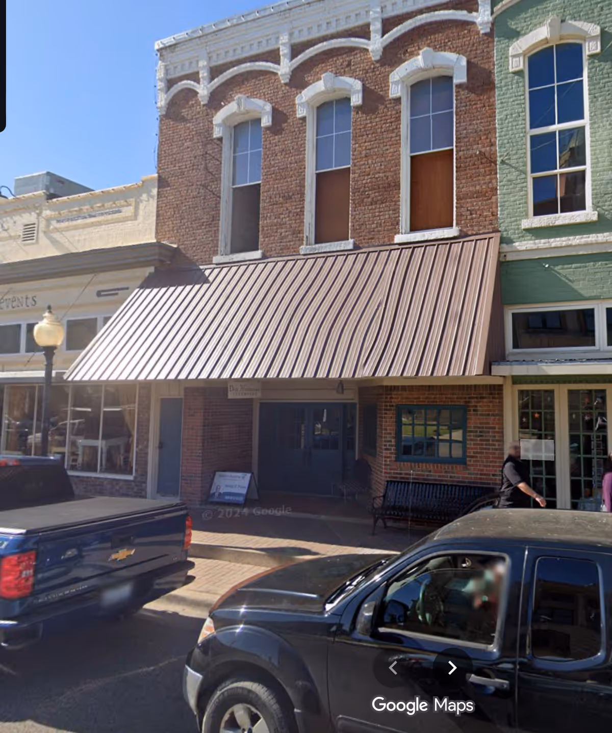 Brick storefront building with brown awning along a sidewalk and parked cars.