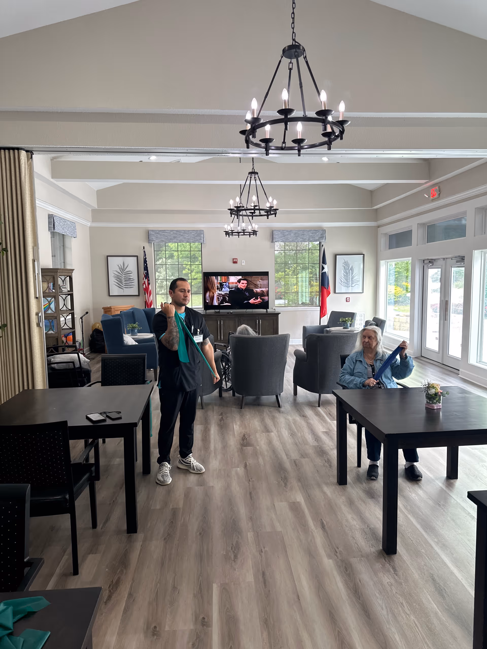Staff member standing near a resident seated at a table in a bright senior living lounge.