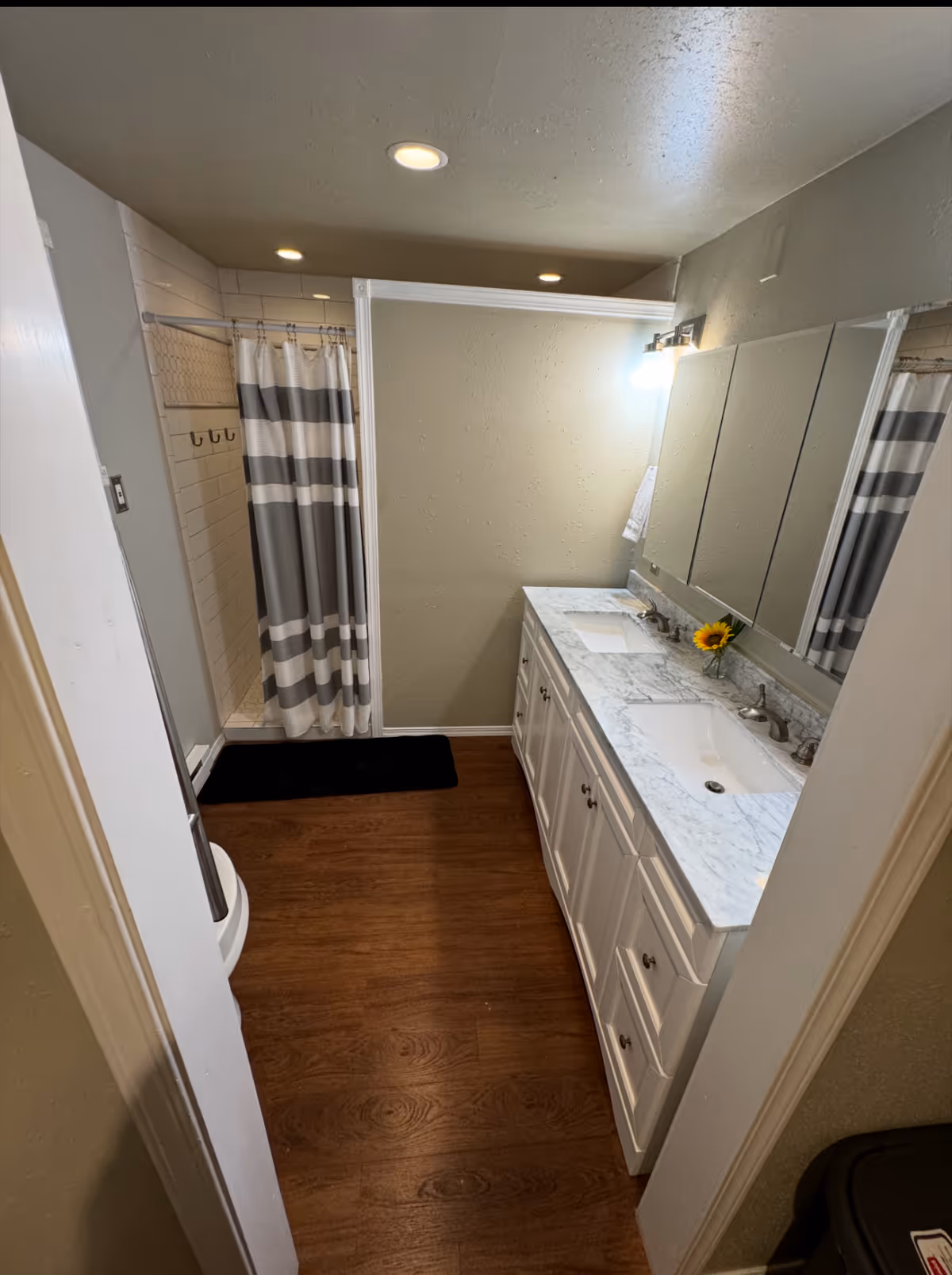 Compact bathroom with dual sinks, marble countertop, shower, striped curtain, and a sunflower.