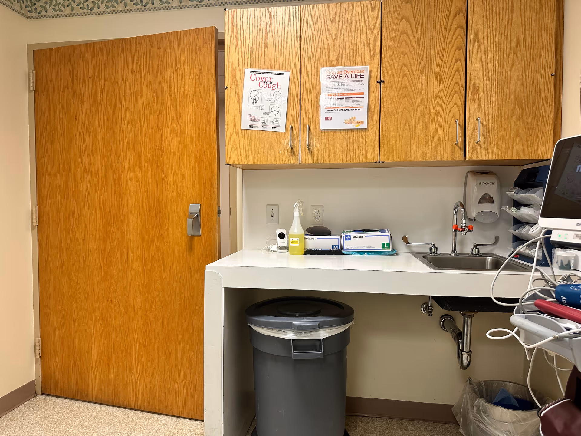 A medical supply area with wooden cabinets, a sink and a trash can